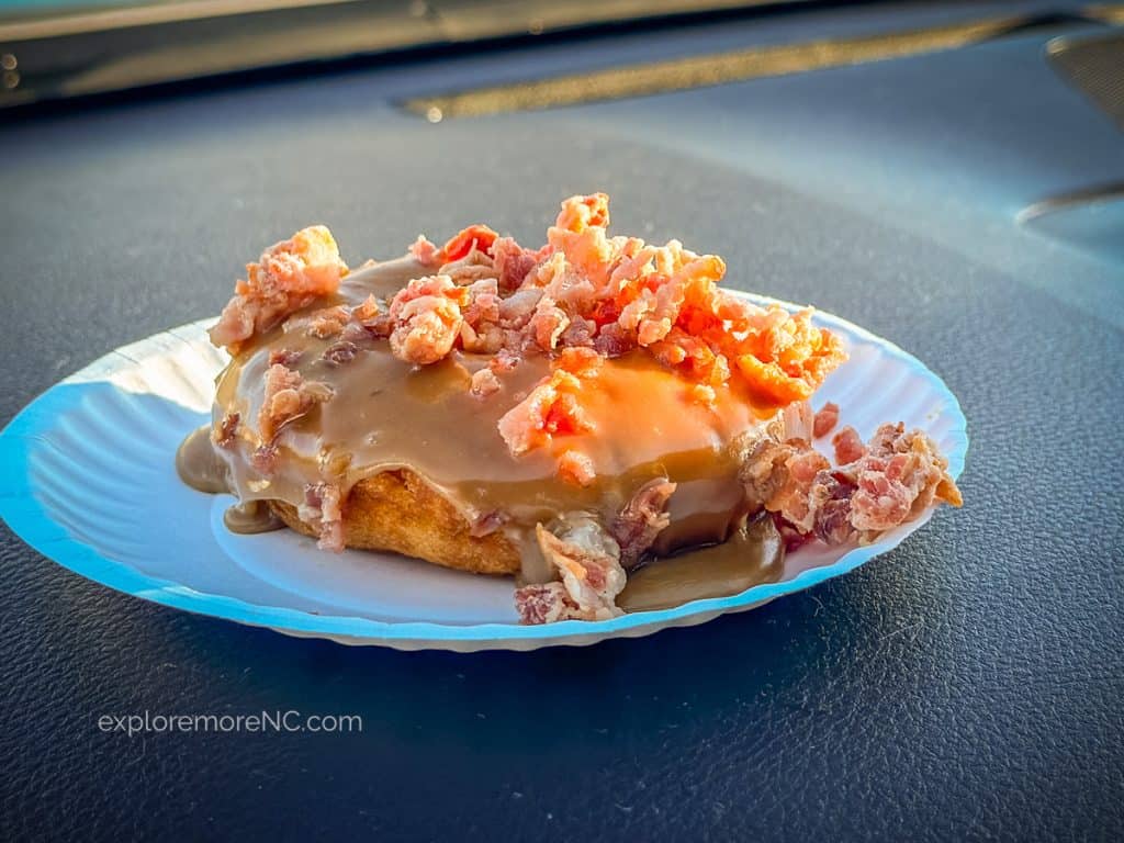 A maple bacon donut with a luscious maple glaze and topped with crunchy pieces of bacon, presented on a paper plate.