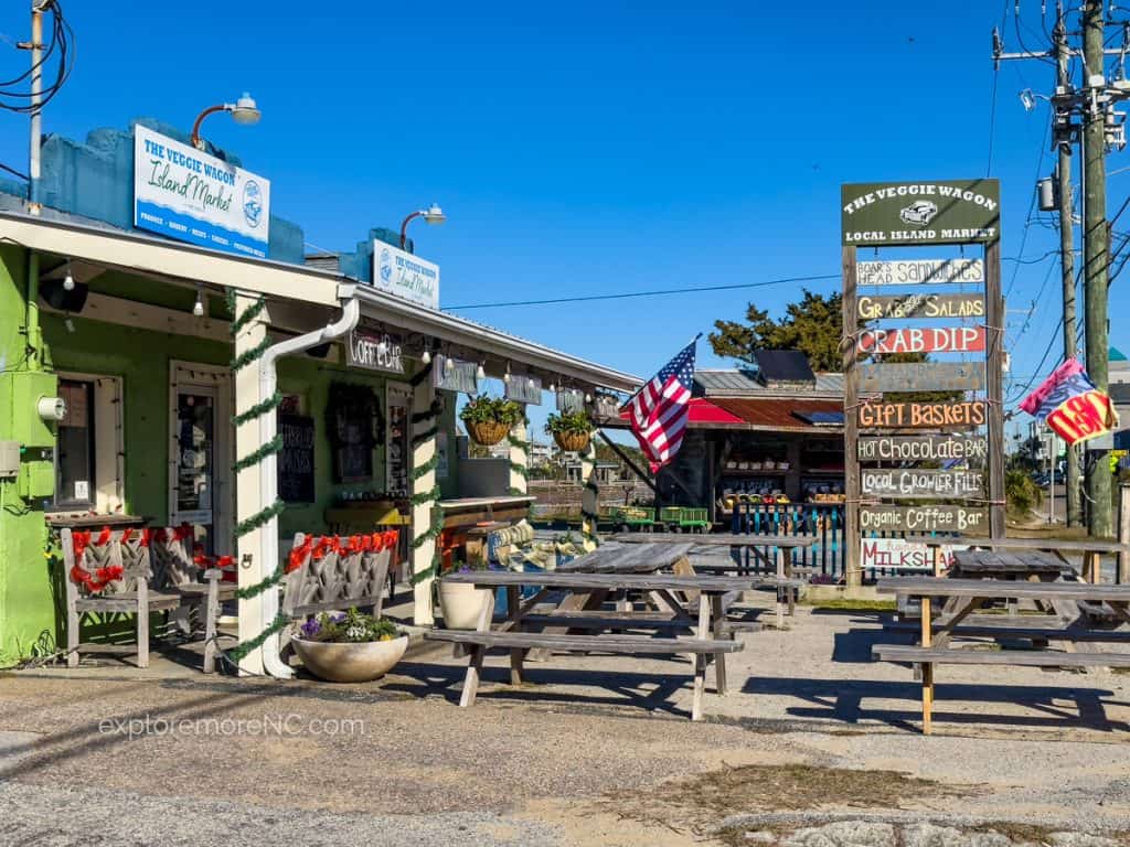 "The Veggie Wagon" local island market, a green-painted store offering various local goods under a clear sky. American flags and signage indicate a friendly, local business atmosphere.